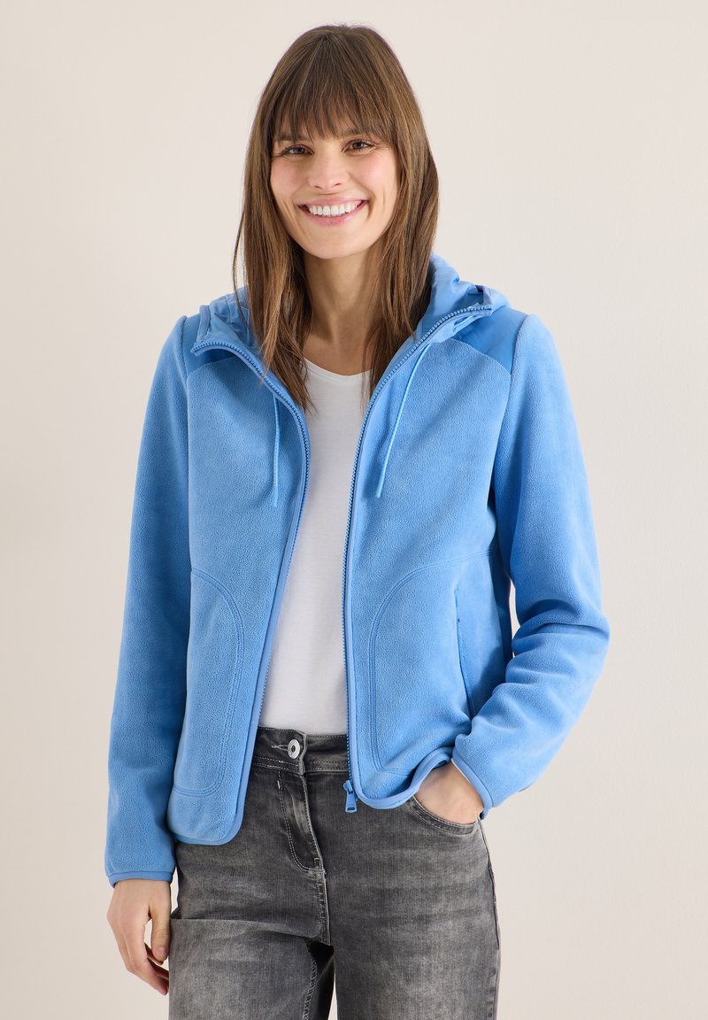 Femme souriante portant une veste en polaire bleu clair avec capuche, chemise blanche et jean gris délavé, se tenant devant un fond uni.