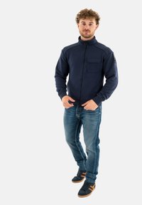 Giacca in pile con zip blu navy e colletto alto, tasca frontale e texture morbida, abbinata a jeans azzurri e sneaker scure.