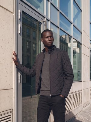 Jeune homme portant une veste matelassée noire et un pantalon foncé, debout devant un bâtiment moderne en verre, appuyant une main contre le mur.