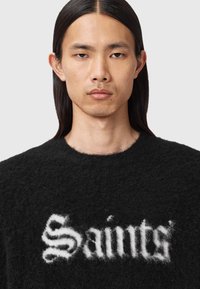 Zwarte donzige trui met het woord "Saints" in witte gestileerde tekst over de voorkant. Ronde halslijn en zachte textuur.