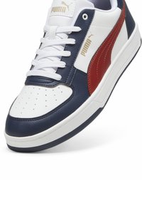 Zapatilla blanca y marina con un acento rojo, presenta una parte superior de cuero suave, caja de dedos perforada y panel lateral texturizado. Logo de PUMA visible.