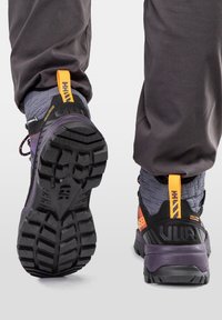 Scarpe da trekking viola e grigie con tomaia texturizzata, suola robusta nera e linguetta arancione sul tallone. Presentano un sistema di allacciatura visibile e dettagli del logo.