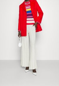 Röd ullkappa, flerfärgad randig stickad tröja, vita texturerade utsvängda byxor. Vit handväska och spetsskor i vitt fulländar looken.