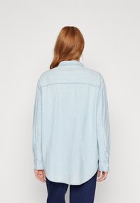 Camicia di jeans azzurro chiaro con colletto, polsini con bottoni e orlo curvo. Presenta dettagli sulla cucitura centrale posteriore e una texture morbida.