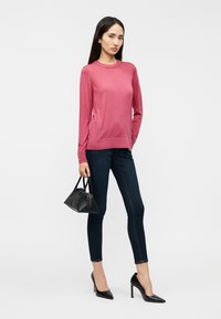 Ροζ πουλόβερ με μακριά μανίκια και στρογγυλή λαιμόκοψη, με λεπτομέρειες από κουμπιά στα μανίκια, συνδυασμένο με σκούρο στενό τζιν και μαύρη τσάντα χειρός.