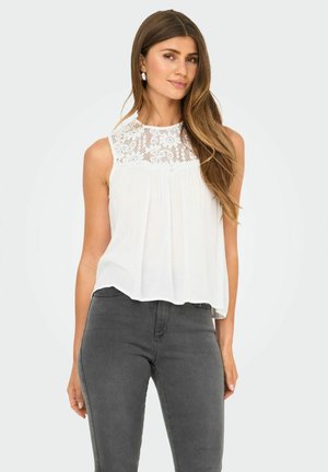 Blusa bianca senza maniche con un gorgiera in pizzo, fronte plissettato e orlo leggermente curvo, abbinata a jeans aderenti grigio scuro.