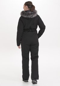Svart isolerad jumpsuit med fuskpälskrage, elastiskt midjeband och sidodragkedjor, designad för vinterförhållanden.