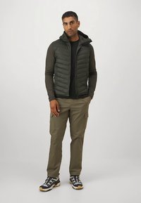 Muška lagana prsluk s kapuljačom tamno zelene boje, uparena s crnom majicom dugih rukava i maslinasto zelenim cargo hlačama s više džepova.