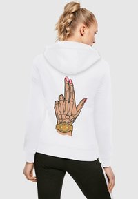 Hvit hettegenser med grafikk av en skjelettfinger med rosa negler og et gullur, vist fra baksiden. Sorte bukser er påført under.