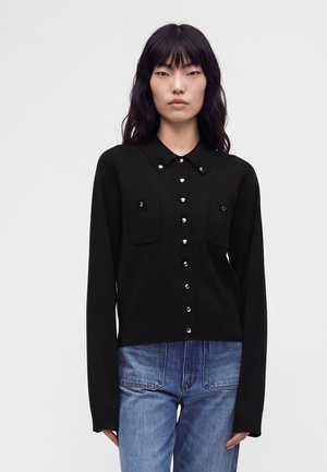 Kvinde med langt sort hår bærer en sort skjorte med knapper og forlommer samt blå jeans mod en ensfarvet lys baggrund.
