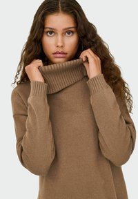Pull à col roulé marron avec col et poignets côtelés, doté d'une texture douce et d'une coupe oversize, tenu près du visage.