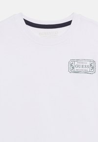 Λευκό μπλουζάκι με στρογγυλή λαιμόκοψη, με μαύρη λεπτομέρεια στο εσωτερικό γιακά και μικρό λογότυπο "GUESS Finest Clothing Company" σε μαύρο, στο πάνω αριστερό μέρος του στήθους.