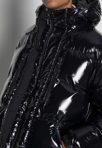 Giacca puffer nera lucida con colletto alto, tasche con zip e un bordo testurizzato a contrasto. Presenta una superficie elegante e lucida.
