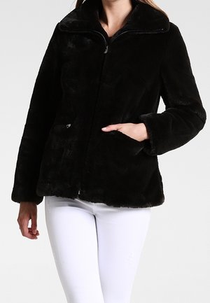 Femme portant une veste zippée en fausse fourrure noire avec col et poches latérales, associée à un pantalon blanc ajusté.