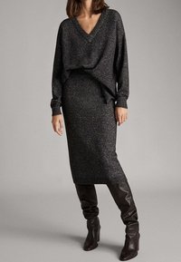 Schwarzer Strickpullover mit V-Ausschnitt und langen Ärmeln, kombiniert mit einem figurbetonten schwarzen Rock. Das Outfit wird durch kniehohe, strukturierte braune Stiefel komplettiert.
