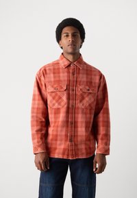 Camisa de cuadro rojo y naranja hecha de algodón, con frente abotonado, dos bolsillos en el pecho y un cuello clásico. Tejido texturizado.