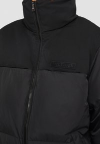 Černá vyp padded bunda s vysokým límcem a předním zipem, s malou obdélníkovou nášivkou loga Hilfiger na prsou.