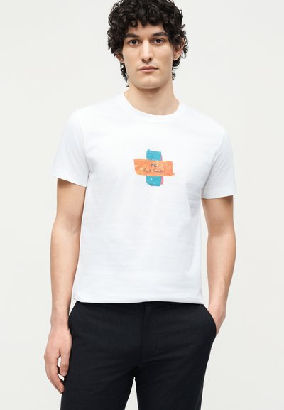 Νέος άνδρας με σγουρά μαλλιά, που φοράει άσπρο μπλουζάκι με μπλε και πορτοκαλί αφηρημένο λογότυπο με την ένδειξη "PS Paul Smith" και μαύρο παντελόνι.