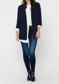 Námořnický blazer s přiléhavým střihem, kombinovaný s bílou košili, tmavě modrými skinny džíny a černými kotníkovými botami. Hladká tkanina a strukturovaný design.