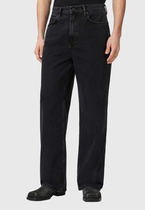 Zwarte jeans met wijde pijpen, gedragen met zwarte schoenen en een zwart topje ingestopt, te zien van taille tot voeten op een effen achtergrond.