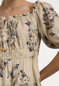 Beige Off-Shoulder-Bluse mit floralem Druck in Blau und Senfgelb, elastischem Taillenbund, kurzen Puffärmeln und gebundenem Ausschnitt mit Holzperlen.