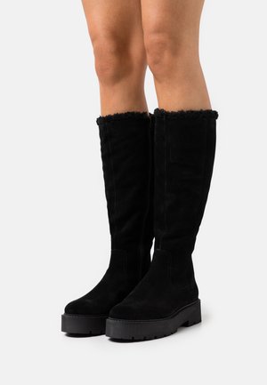 Bottes noires montant jusqu'aux genoux avec semelles compensées épaisses et bordure en fourrure au sommet, portées sur des jambes nues sur un fond blanc.
