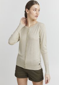 Lyse beige cardigan laget av mykt strikket stoff med rund hals, front knappelukking og ribbet kant, kombinert med olivengrønne shorts.