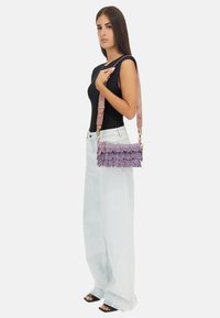 Borsa viola con frange e tracolla rosa. Presenta strati testurizzati e accessori in oro. Il modello indossa una maglietta nera e jeans a gamba larga in denim chiaro.