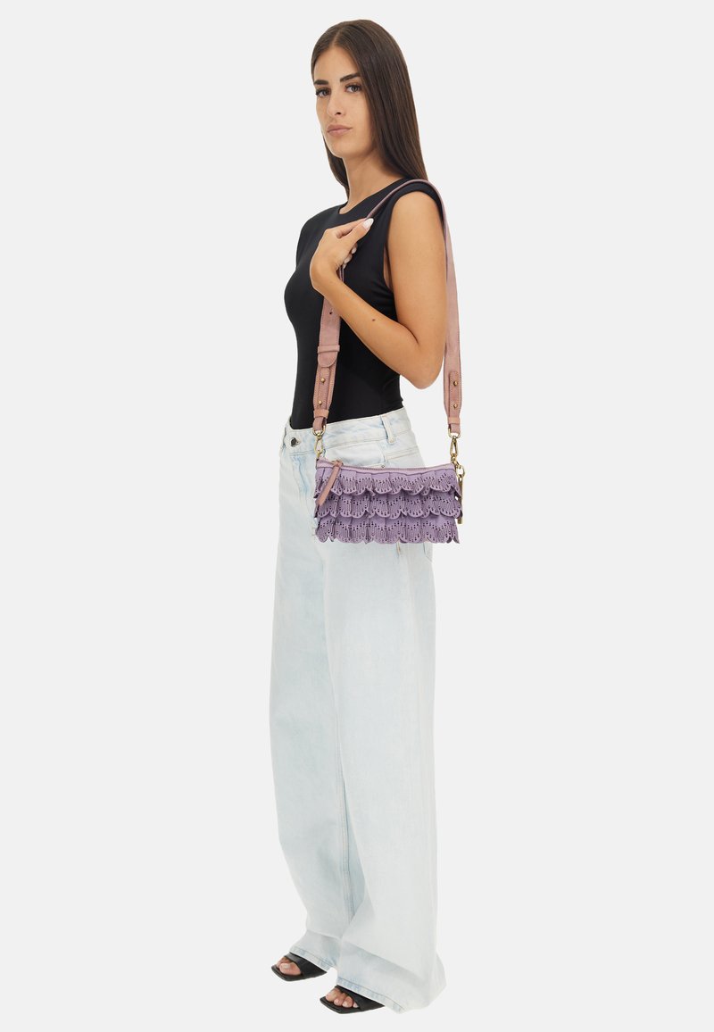 Borsa viola con frange e tracolla rosa. Presenta strati testurizzati e accessori in oro. Il modello indossa una maglietta nera e jeans a gamba larga in denim chiaro.