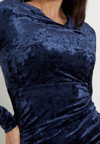 Mörkblå sammetsklänning med en texturerad yta, med en omslagdesign och långa ärmar. Tyget verkar mjukt och lyxigt.