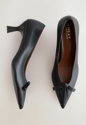 Pantofi cu toc înalt din piele neagră, cu vârf ascuțit și un mic accent sub formă de fundiță în față. Textură netedă și design elegant.