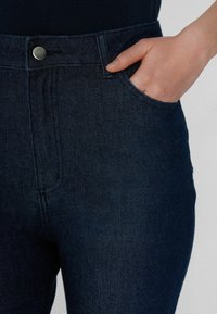 Jeans de mezclilla azul oscuro con una cintura alta, que cuentan con un bolsillo delantero y un acento de botón metálico. Textura suave y diseño ajustado.