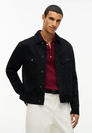 Jeune homme portant une veste noire boutonnée sur un polo rouge et un pantalon blanc, debout devant un fond blanc uni.