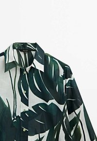 Chemise à col à manches longues avec un grand imprimé de feuilles tropicales vert foncé et noires sur un tissu blanc transparent, boutonnée à l'avant.