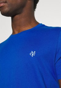 Königsblaues T-Shirt mit kurzen Ärmeln aus Baumwollmischung, mit einem weißen Logo auf der linken Brustseite und einem Rundhalsausschnitt. Glatte Textur.