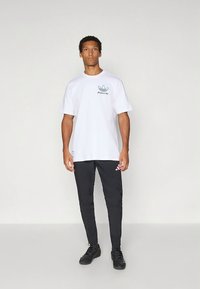 Balti trumpomis rankovėmis marškinėliai su žalia grafine logotipo ant krūtinės, derinami su juodomis siaurėjančiomis kelnėmis su rožiniu prekės ženklo akcentu kairėje kojoje. Juodi batai.