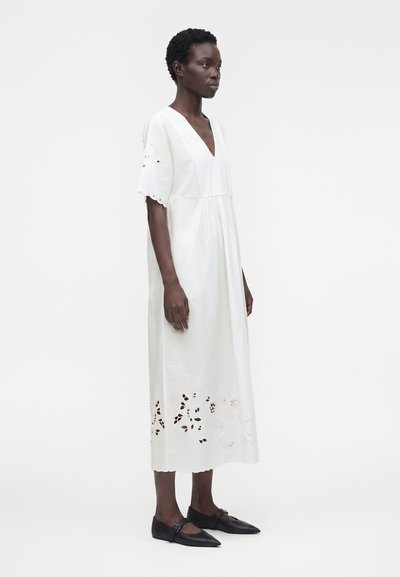 Femme debout dans une robe midi blanche à manches courtes avec des détails découpés floraux près de l'ourlet, portant des chaussures plates noires.