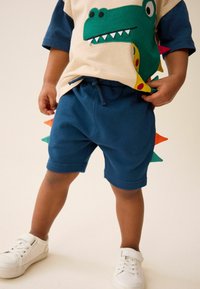 Children's outfit featuring a beige shirt with green dinosaur graphic and blue shorts with colorful spikes; white sneakers complete the look.