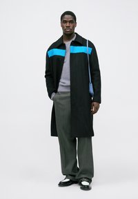 Palton lung negru cu o dâră albastră pe piept, purtat peste un pulover gri și pantaloni gri. Accesoriile includ o geantă mică albastră și pantofi negri.