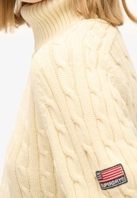 Maglione a collo alto a coste color crema con motivo a trecce. Presenta un patch con il logo dettagliato con la bandiera americana sulla manica.