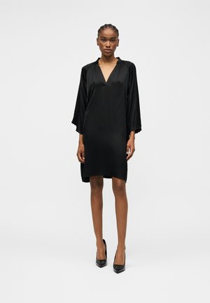 Kvinde iført en knælange sort satin kjole med brede ærmer og sorte høje hæle, stående mod en ensfarvet lys baggrund.