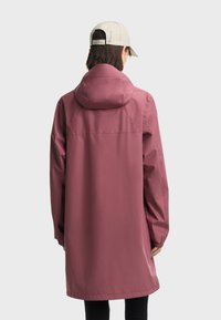 Osoba u dugom ljubičasto-sivom kišnom kaputu s kapuljačom i bež kapom, okrenuta leđima prema jednostavnoj svijetloj pozadini.