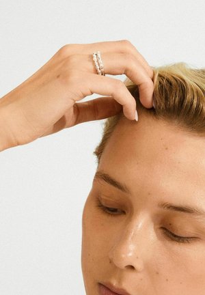Primo piano di una persona che tocca i propri capelli biondi corti, indossando due anelli di perline argentate sull'indice, su uno sfondo bianco.