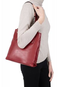 Borsa tote rossa in pelle con texture liscia, due tracolle e design minimalista, portata da una persona che indossa un maglione beige.