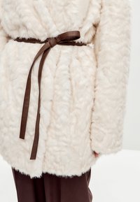Faux fur frakke i off-white med en textured finish, belagt med et mørkebrunt læderbælte i taljen. Klassisk skæring med brede ærmer.