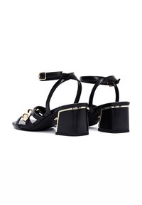 Sandalias de tacón de cuero negro con correas en el tobillo ajustables, acentos dorados y tacones en bloque gruesos con un acabado texturizado.