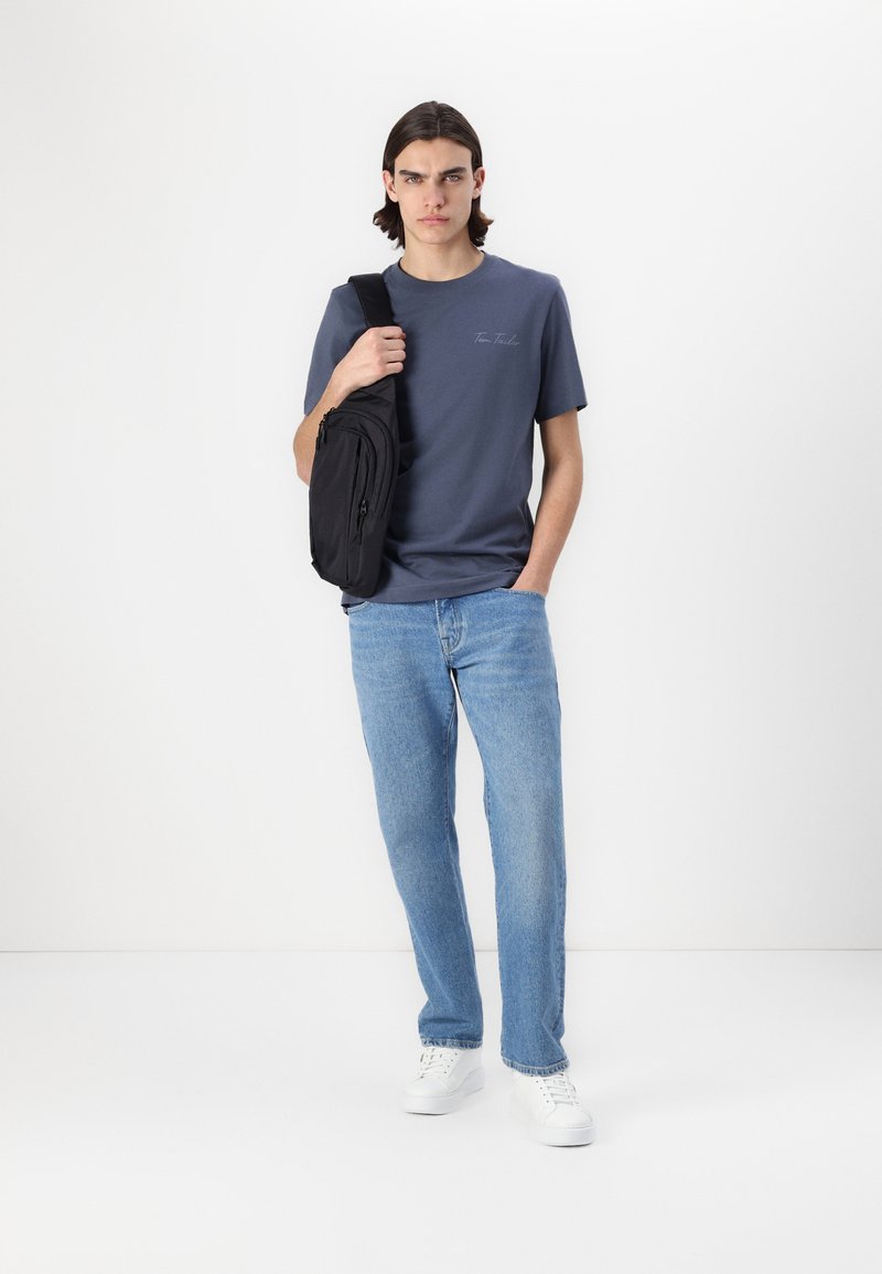 Marineblå bomulls t-skjorte med logodetalj, paret med lyseblå jeans og hvite sneakers. En svart crossover-veske henger over skulderen.