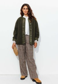 Veste moelleuse vert olive avec fermeture à bouton, blouse texturée blanche, pantalon large à carreaux et bottes en daim marron. Les accessoires incluent une pochette.