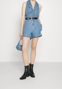 Denimový overal světle modré barvy s límečkem, knoflíky, dvěma bočními kapsami a upnutým černým páskem. Kombinovaný s černými kotníkovými botami.