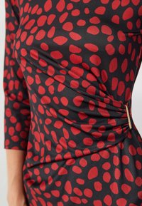 Schwarzes Kleid mit roten unregelmäßigen Punkten, das ein schmeichelhaftes Raffungdesign und einen goldenen Akzent an der Taille aufweist. Glattes, dehnbares Material.